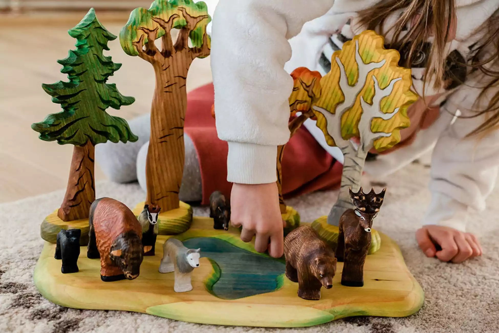A child playing with a wooden forest playset featuring trees, a pond, and various animal figures including bears, a deer, and a wolf.